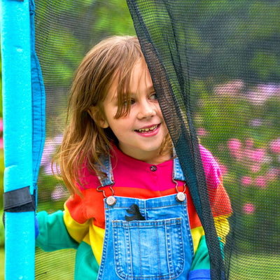 A child in denim overalls and a rainbow-striped shirt stands beside the TP Toys TP UP 6ft Trampoline outdoors, showcasing its fun and safety features for kids amid green grass and pink flowers.