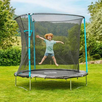 A red-haired child jumps on a TP Toys TP Up 8ft Trampoline with zip-free entry, set on a grassy lawn with trees and hills in the background.