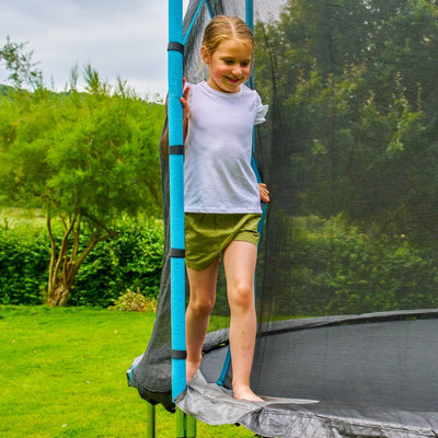 A young girl in a white shirt and green shorts steps onto a TP Toys TP Up 8ft Trampoline outdoors, surrounded by green grass and trees.