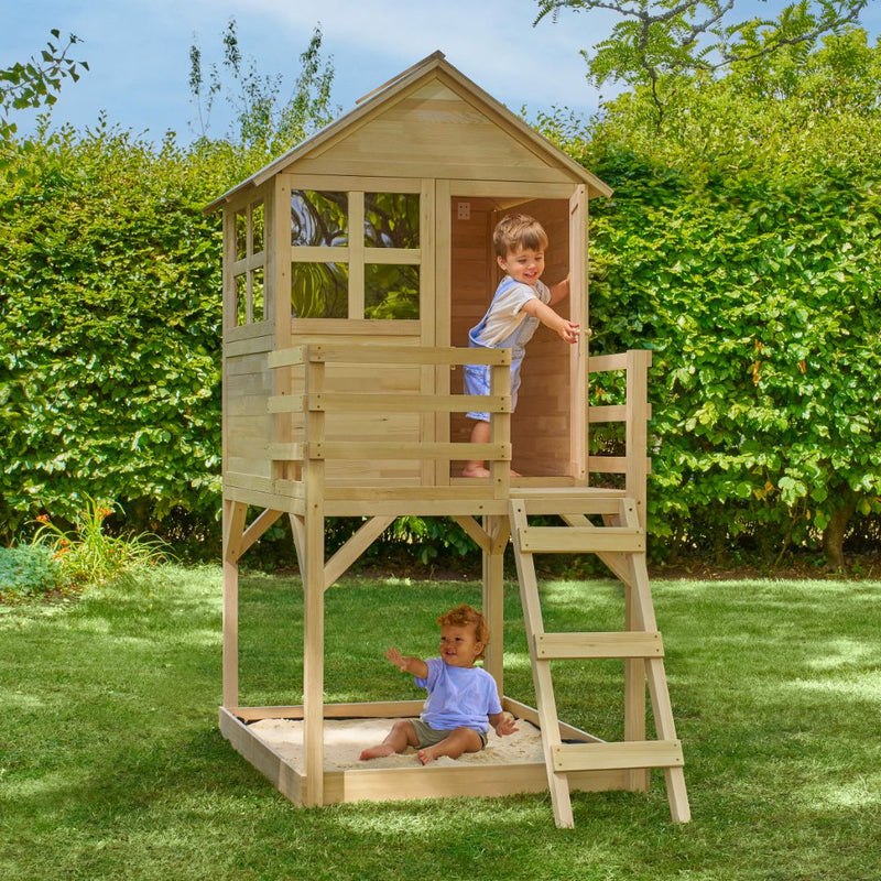 Two young children play on the TP Toys Buttercup Cottage Wooden Playhouse with Deck—made from FSC® certified wood—which features a ladder and sandbox in a grassy backyard surrounded by green bushes.