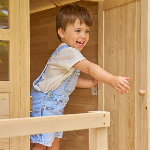 A young child smiles while holding the door of the TP Toys Buttercup Cottage Wooden Playhouse with Deck, made from FSC® certified wood.
