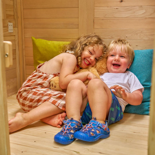 Two young children smile and hug a stuffed animal on the floor of the TP Toys Buttercup Cottage Wooden Playhouse with Verandah, 6ft Wavy Slide, Double Swing Arm, and Deck—creating a cozy space with colorful pillows behind them.