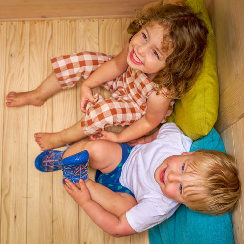 Two young children smile and look up at the camera as they sit on colorful cushions inside the TP Toys Buttercup Cottage Wooden Playhouse with Deck, Double Swing Arm & 6ft Wavy Slide – FSC® certified.