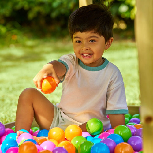 A young child smiles while sitting in a ball pit outdoors next to the TP Toys Buttercup Cottage Wooden Playhouse with Deck & 6ft Wavy Slide, surrounded by colorful balls and lush greenery.