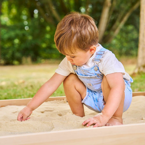 A young child wearing denim overalls and a white shirt plays in the sand near the TP Toys Buttercup Cottage Wooden Playhouse with Deck & 6ft Wavy Slide (FSC® certified), set outdoors amid grass and trees.