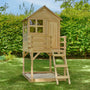 The TP Toys Buttercup Cottage Wooden Playhouse with Deck stands elevated on stilts in a grassy backyard, made from FSC® certified wood, featuring windows, a small porch, a ladder, and a sand area below.