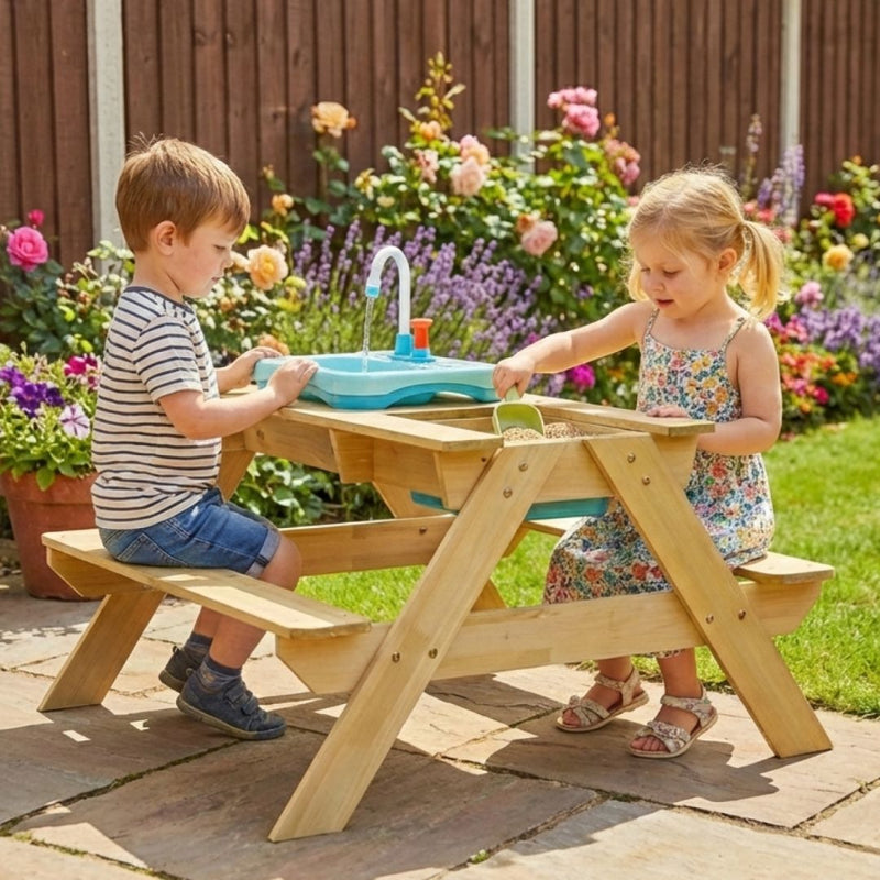 Two young children enjoy the TP Toys TP Splash & Play Wooden Picnic Table, featuring a built-in play sink and sand area, while sitting together in a flower-filled garden.