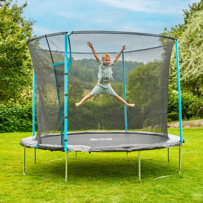 A child jumps with arms and legs spread on the TP Toys TP Up 10ft Trampoline, featuring protective bounce pads and safety netting, in a grassy backyard.