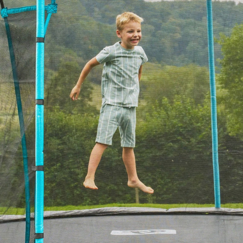 A young boy leaps on the TP Toys TP Up 10ft Trampoline, which features protective bounce pads and a safety net, surrounded by trees and hills in the background.