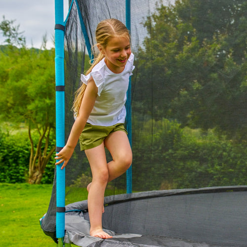 A young girl with blonde hair, in a white shirt and green shorts, stands outdoors on a TP Toys TP Up 12ft Trampoline, holding the safety net pole.