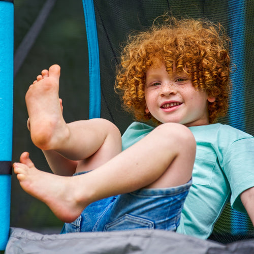 A young child with curly red hair and freckles smiles outdoors, bare feet up on the TP Toys TP Up 12ft Trampoline, wearing a light blue shirt and denim shorts.