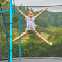 A young girl in a white shirt and green shorts jumps with arms and legs outstretched on the TP Toys TP Up 12ft Trampoline outdoors, surrounded by trees and grass.