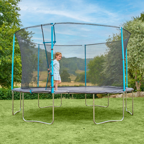 A young child stands on the TP Toys TP Up 14ft Trampoline with a safety net enclosure in a grassy backyard on a clear day.