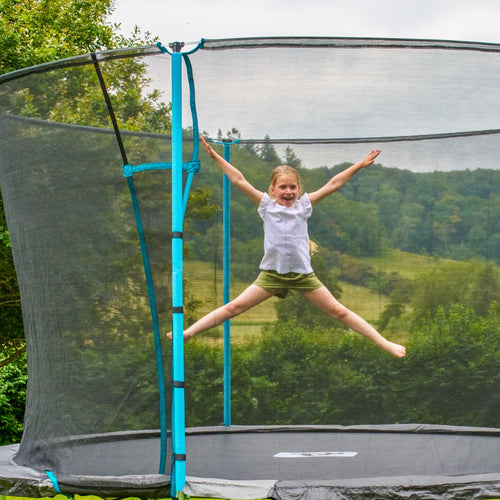A young girl jumps with arms and legs outstretched on the TP Toys TP Up 14ft Trampoline, safely enclosed by a net, with trees and hills visible in the background.