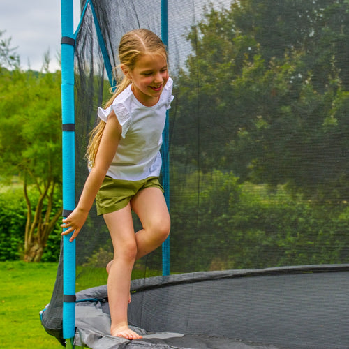A blonde-haired girl in a ponytail smiles as she stands on a TP Up 14ft Trampoline by TP Toys outdoors, holding the safety net and wearing a white shirt with green shorts.