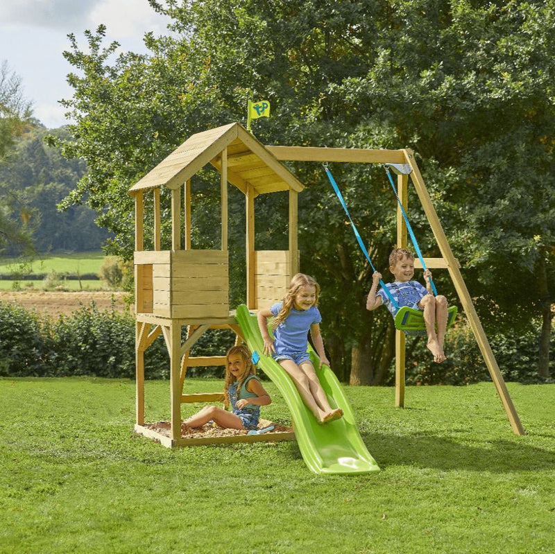 Three children play on the TP Toys TP Treehouse Wooden Play Tower with Wavy Slide & Swing Arm (FSC® certified), which includes a swing and sandbox, set outdoors on grass with trees in the background.