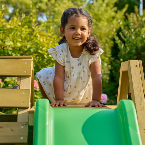 A young girl smiles as she kneels at the top of the green slide on the TP Wooden Toddler Climb & Slide by TP Toys, an FSC® certified outdoor play structure.