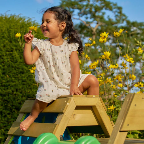 A young girl in a light floral dress smiles as she sits on the TP Toys TP Wooden Toddler Climb & Slide - FSC® certified, surrounded by yellow flowers and greenery outdoors under a clear blue sky.