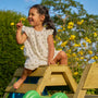 A young girl in a light floral dress smiles as she sits on the TP Toys TP Wooden Toddler Climb & Slide - FSC® certified, surrounded by yellow flowers and greenery outdoors under a clear blue sky.