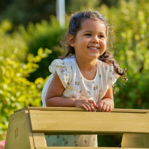 A young girl in a floral dress smiles behind the TP Toys TP Wooden Toddler Climb & Slide—FSC® certified—ready for active play in a sunlit garden.