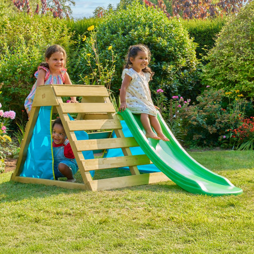 Three young children enjoy active play on the TP Toys TP Wooden Toddler Climb & Slide – FSC® certified, set up in a grassy garden surrounded by flowering plants.