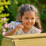 A young girl with curly hair smiles as she leans over the TP Toys TP Wooden Toddler Climb & Slide - FSC® certified, enjoying outdoor play surrounded by greenery and flowers.