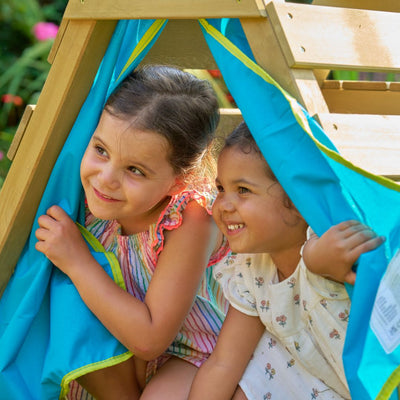 Two young girls smile and peek out from their hiding spot under the TP Toys TP Wooden Toddler Climb & Slide – FSC® certified, enjoying active play beneath its blue fabric.
