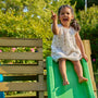 A young girl enjoys active play on the TP Wooden Toddler Climb & Slide by TP Toys, made with FSC® certified wood, smiling and pointing upward amid trees and flowers outdoors.