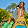 A young girl smiles atop the TP Toys TP Wooden Toddler Climb & Slide - FSC® certified, enjoying active play outdoors surrounded by green plants and yellow flowers.