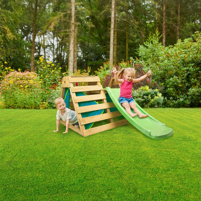 Two young children play energetically on the TP Toys TP Wooden Toddler Climb & Slide - FSC® certified, featuring a green slide, in a grassy backyard bordered by trees and bushes.