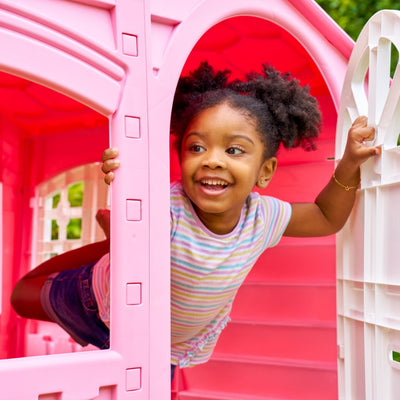 A young girl in a striped shirt and denim shorts smiles as she leans out of the TP Toys Palplay Plastic Playhouse of Fun - Candy Floss Edition.