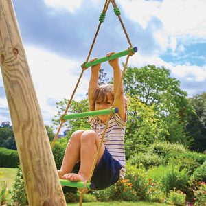 A child climbs the rope ladder of the TP Knightswood Double Wooden Swing Set With Glide Ride & Rope Ladder by TP Toys, which is crafted from PEFC certified wood and set outdoors among greenery under a blue sky.