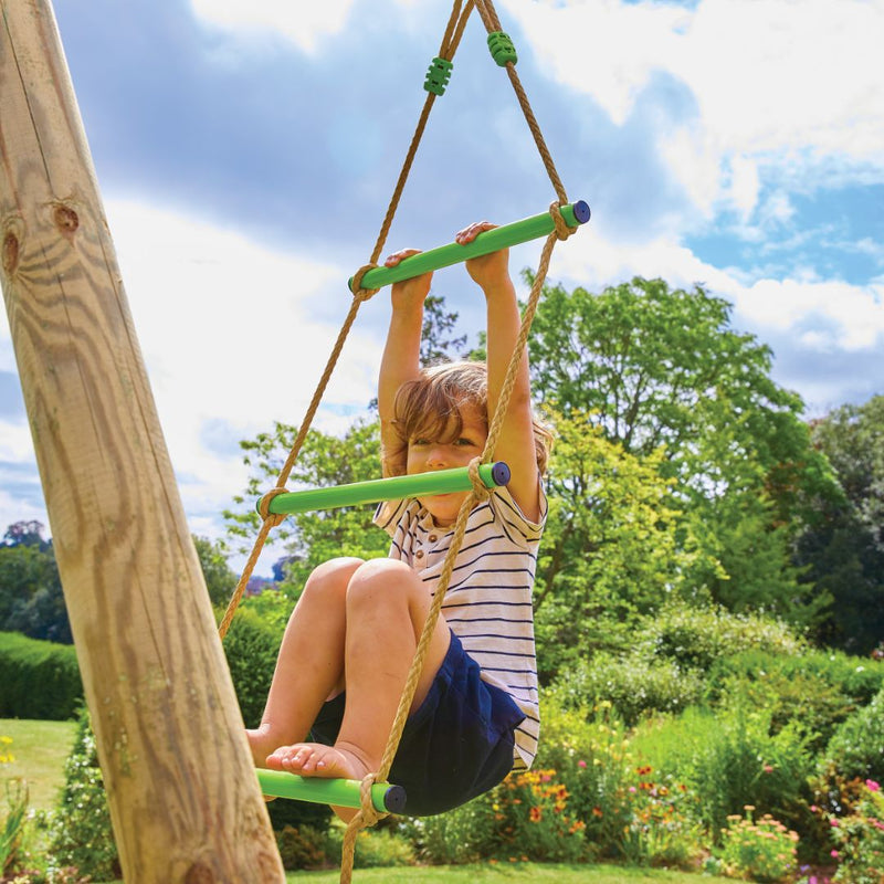 A child climbs the rope ladder of the TP Knightswood Double Wooden Swing Set With Glide Ride & Rope Ladder by TP Toys, which is crafted from PEFC certified wood and set outdoors among greenery under a blue sky.