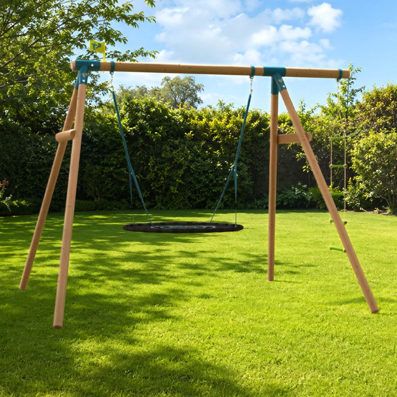The TP Toys TP Knightswood Double Wooden Swing Set With Giant Nest Swing & Rope Ladder – PEFC Certified features a round black giant nest swing hanging from its sturdy wooden frame, set on a green lawn in a backyard surrounded by trees and hedges under a partly cloudy sky.