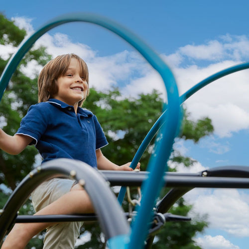 A young boy in a blue polo shirt smiles while climbing the TP UFO Den & Climbing Frame by TP Toys, an exciting piece of garden play equipment outdoors under a partly cloudy sky.