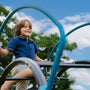A young boy in a blue polo shirt smiles while climbing the TP UFO Den & Climbing Frame by TP Toys, an exciting piece of garden play equipment outdoors under a partly cloudy sky.