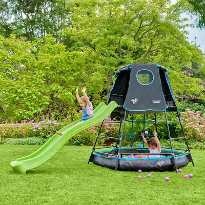 Two children enjoy the TP Toys TP Explorer Metal Climbing Frame Set and Ripple Slide Black Edition, featuring a green slide and ball pit. The sturdy metal frame adds excitement to their backyard playtime, surrounded by lush grass and plants.