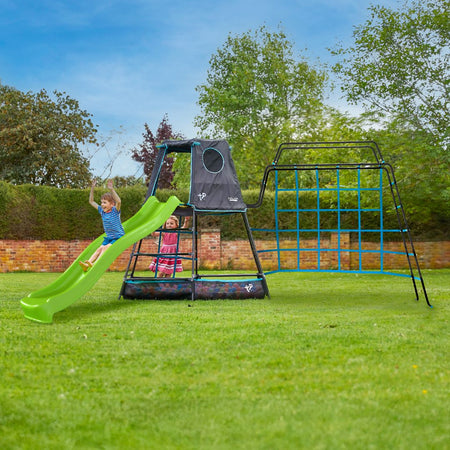 Two young children play on the TP Toys TP Explorer Metal Climbing Frame Set with Monkey Bridge & Ripple Slide Black Edition in a grassy backyard, surrounded by trees, shrubs, and a brick wall on a sunny day.