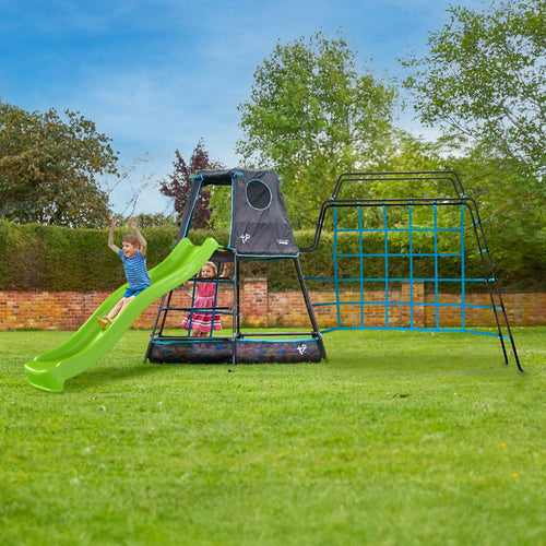 Two young children play on the TP Toys TP Explorer Metal Climbing Frame Set with Monkey Bridge & Ripple Slide Black Edition in a grassy backyard, surrounded by trees, shrubs, and a brick wall on a sunny day.