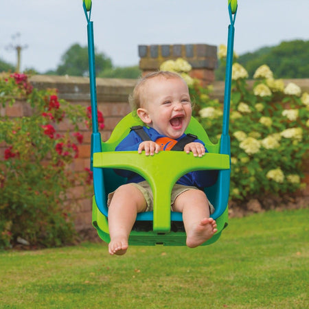 A smiling baby sits in a TP Toys Quadpod on the TP Small to Tall 2 in 1 Metal Swing Set, holding the front bar outdoors with flowers and a brick wall behind.