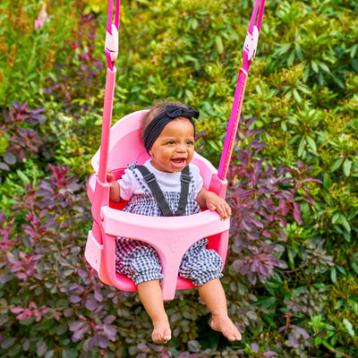 A baby in a headband and checkered overalls smiles while sitting in the pink Quadpod seat of the TP Toys Small to Tall 2 in 1 Metal Swing Set, with green and purple foliage in the background.