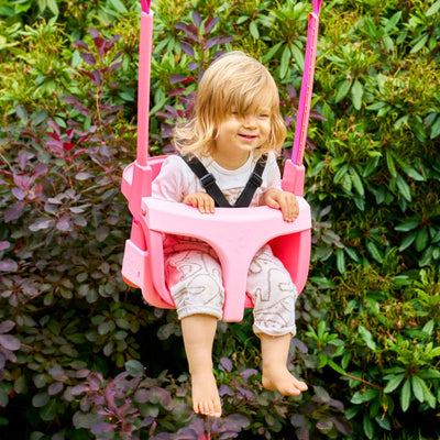 A young blonde child sits barefoot in a pink Quadpod seat on the TP Toys Small to Tall 2 in 1 Metal Swing Set, surrounded by green bushes—an ideal grow-with-me swing for endless outdoor play.