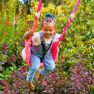 A young child in denim overalls and a blue bow smiles while swinging on the pink Quadpod of the TP Small to Tall 2 in 1 Metal Swing Set by TP Toys.