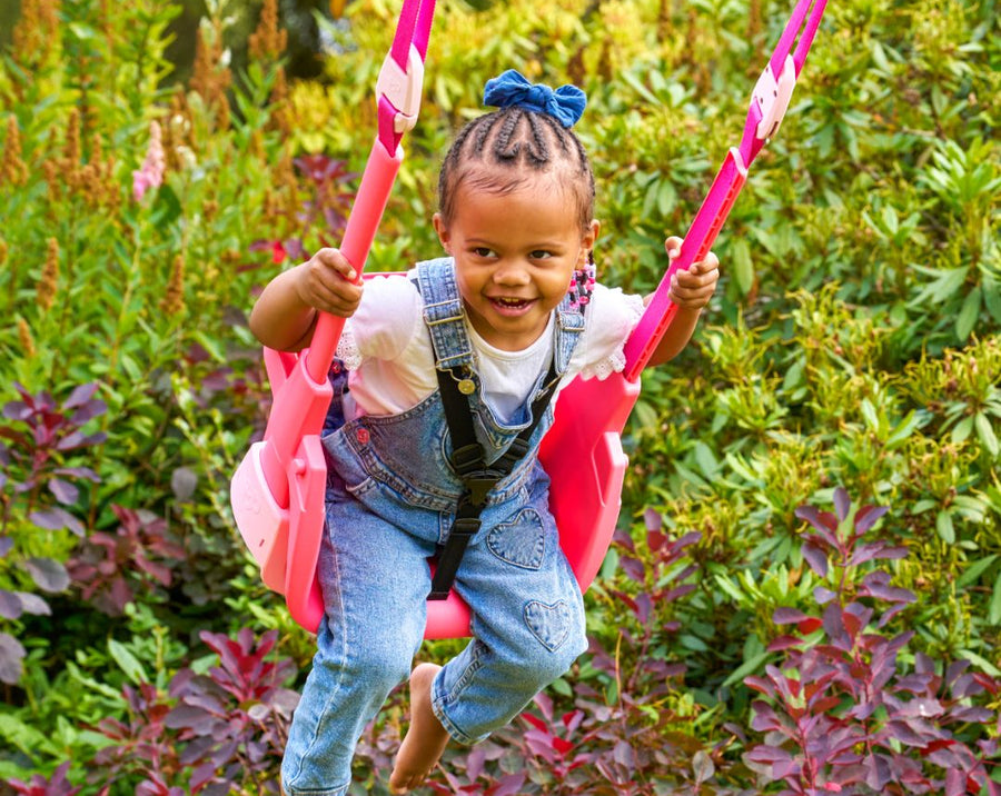 A young child swings outdoors on the TP Toys Forest Acorn Growable Swing Set with Quadpod—FSC® certified—enjoying playtime on a sturdy frame surrounded by green and red foliage.