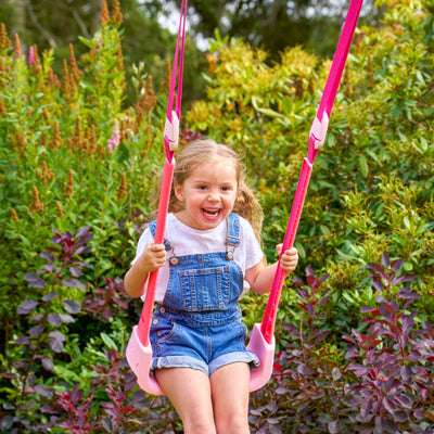 A young girl in denim overalls and a white t-shirt smiles as she swings on the pink Quadpod of the TP Toys Small to Tall 2 in 1 Metal Swing Set, surrounded by green and reddish plants in a garden.