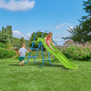 A girl enjoys the TP Toys TP 6ft (1.8m) Wavy Slide & Stepset outdoors while a boy walks nearby on the grassy lawn, surrounded by trees and bushes in a garden playground.
