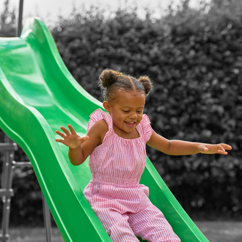A young child with pigtails, dressed in a pink striped outfit, slides down the bright green TP Toys 3m/10ft Super Wavy Slide outdoors, surrounded by greenery.