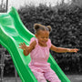 A young child with pigtails, dressed in a pink striped outfit, slides down the bright green TP Toys 3m/10ft Super Wavy Slide outdoors, surrounded by greenery.