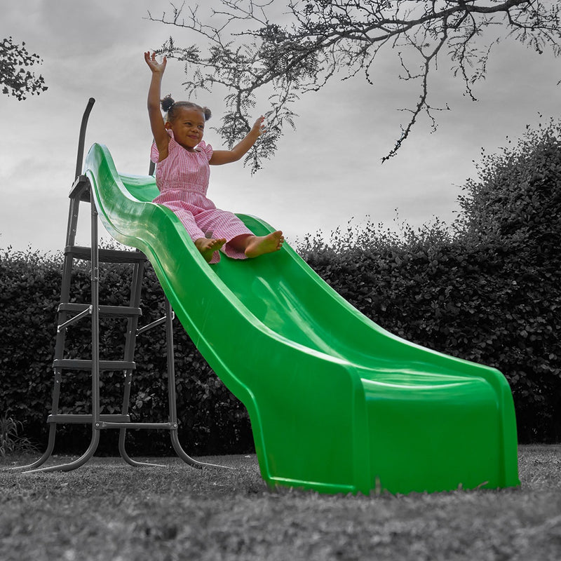 A young child in a pink outfit slides down the bright green TP 3m/10ft Super Wavy Slide by TP Toys, enjoying the UV stabilised outdoor play equipment in a yard surrounded by trees and bushes.