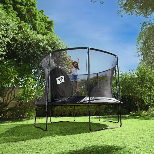 A child jumps on a TP Toys Genius® 14ft Round Trampoline with a black finish, galvanised steel frame, and SurroundSafe enclosure in a sunny backyard surrounded by trees and grass.